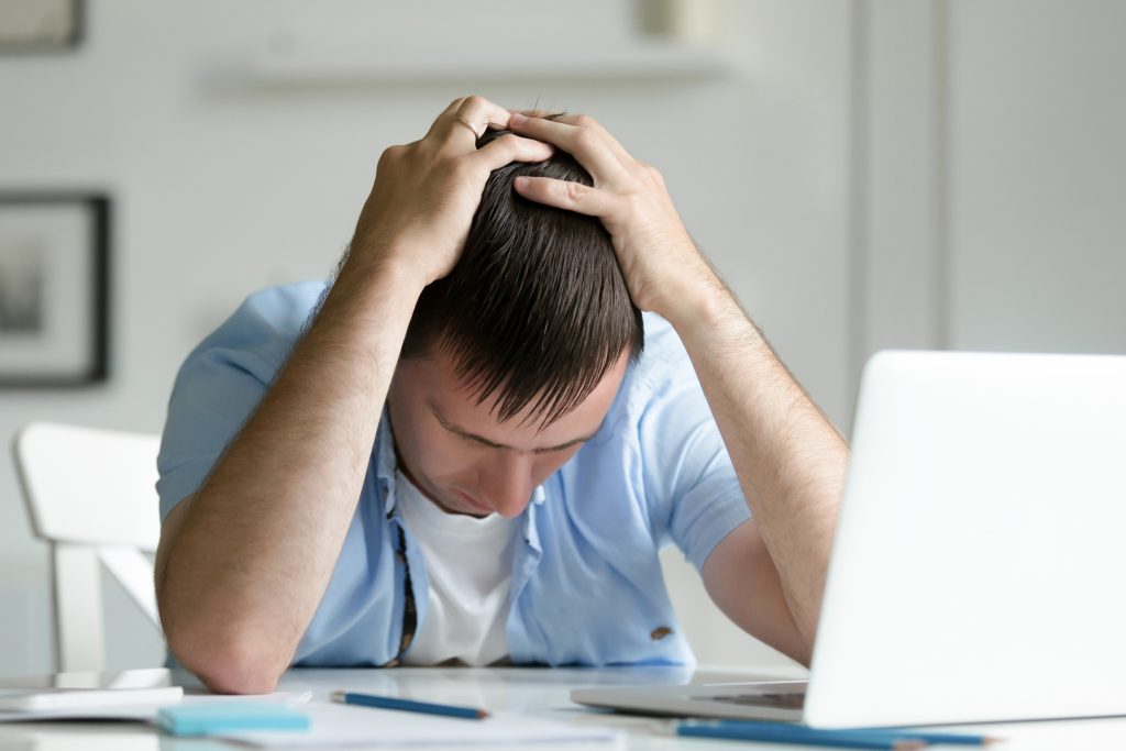 Young man with his head in his hands while seated at a table with a laptop.