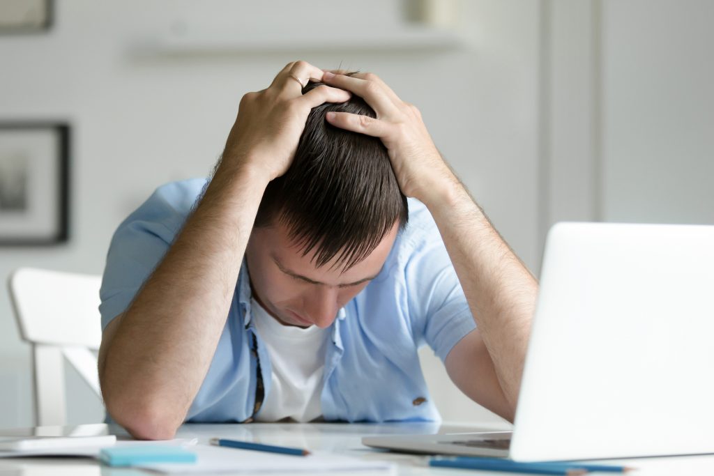 Young man with his head in his hands while seated at a table with a laptop.