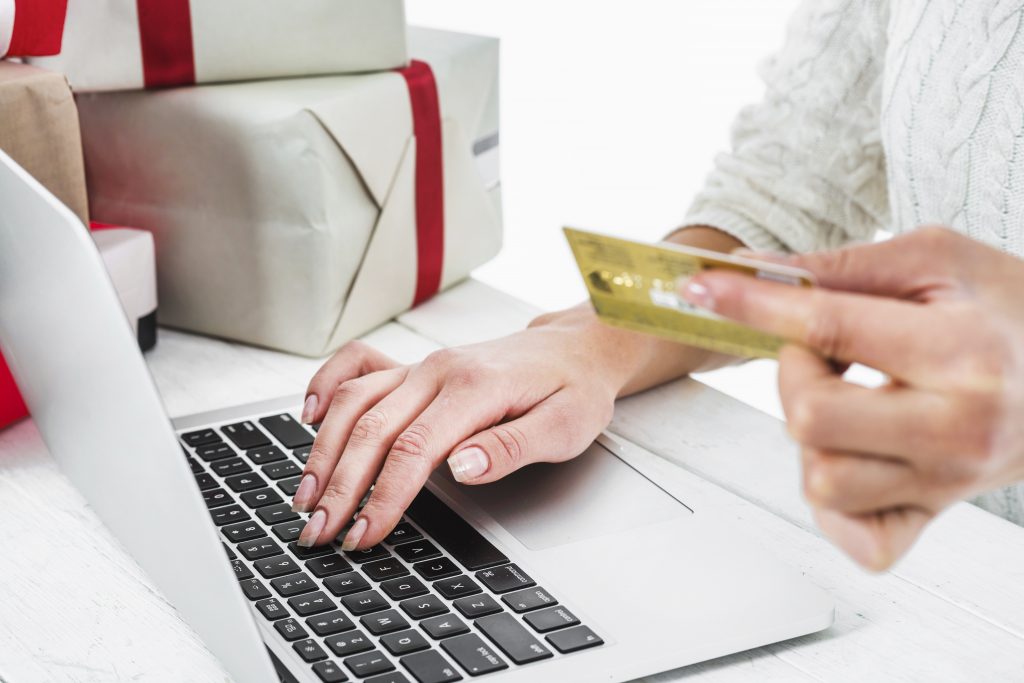 Woman sitting at a laptop with credit card in hand with wrapped holiday presents at her side.