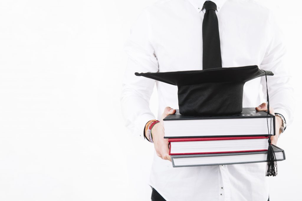 Man in white button up shirt with dark tie carrying textbooks with a graduation cap on the books.