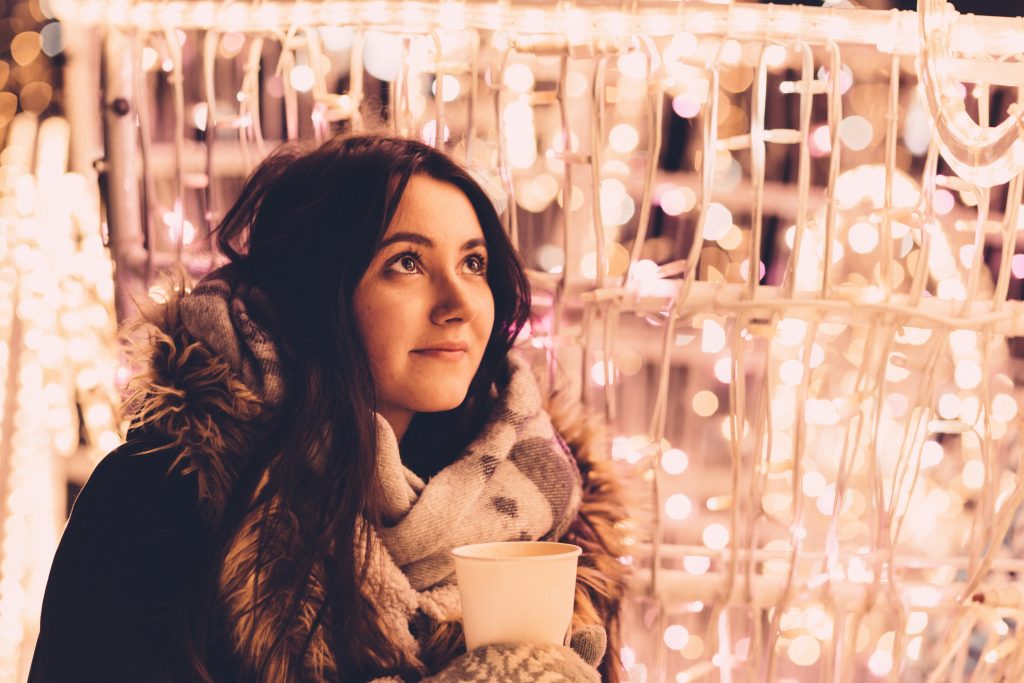 Young girl bundled up in coat, scarf and mittens with a hot drink in front of a wall of white lights.